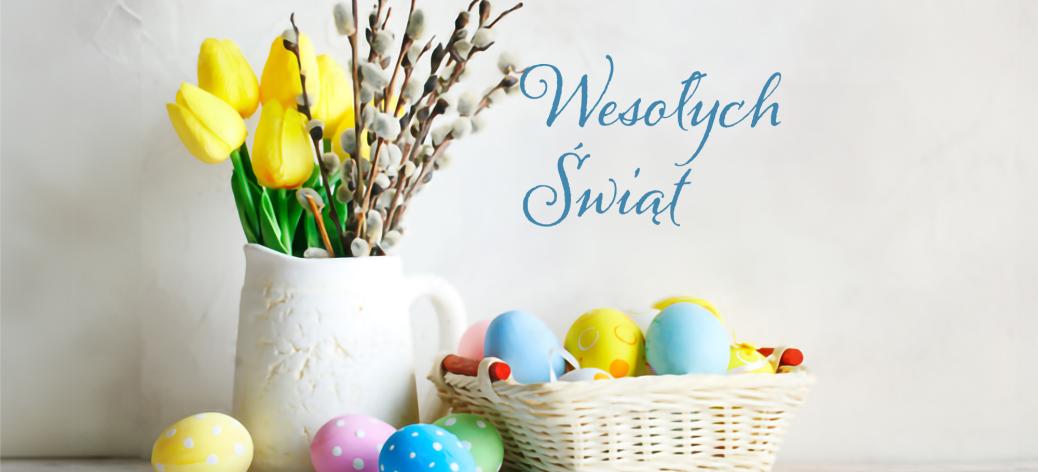 Na zdjęciu napis Wesołych Świąt, dzbanek z żółtymi tulipanami i baziami  obok koszyczek z jajkami oraz luźno leżące różnokolorowe  jajka