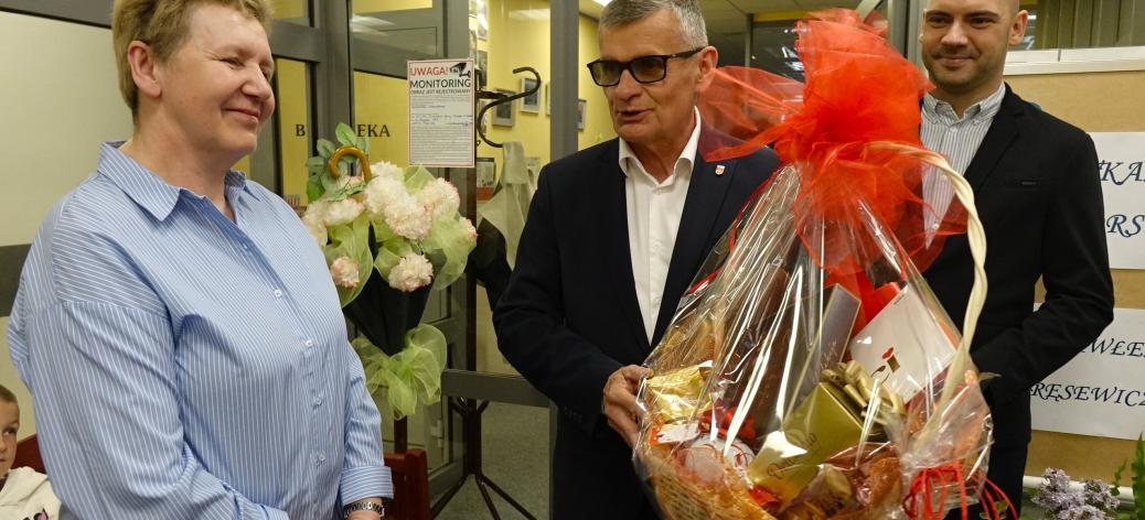 Uroczystość w pomieszczeniu biblioteki. Po lewej stoi uśmiechnięta Dyrektor MBP.  W środku Burmistrz Drzewicy trzymający duży kosz prezentowy ozdobiony czerwonymi kokardami. Obok stoi Zastępca Burmistrza. W tle przeszklone drzwi z napisem „Biblioteka”, kwiaty oraz elementy wystroju wnętrza.