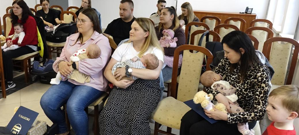 Grupa rodziców z niemowlętami podczas oficjalnego spotkania w Sali USC. W centralnej części siedzą trzy kobiety trzymające na rękach małe dzieci. W tle widoczni są pozostali uczestnicy siedzący na drewnianych krzesłach z jasną tapicerką.