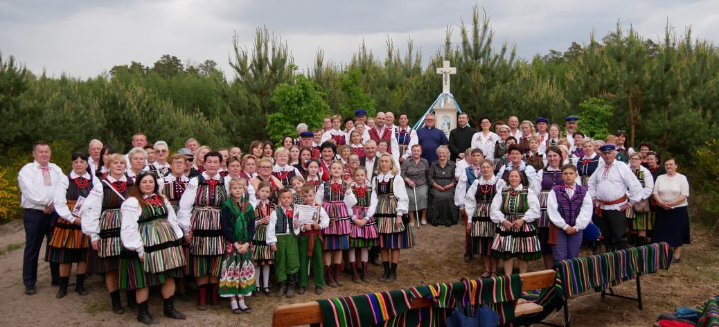 Duża grupa uczestników Majówki na ludowo w Gminie Drzewica, ubrana w barwne, tradycyjne stroje opoczyńskie, pozuje do pamiątkowego zdjęcia przy leśnej kapliczce w towarzystwie dzieci i kapłanów