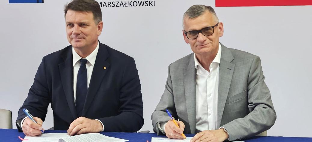 Członek Zarządu Województwa Łódzkiego Artur Bagieński i Burmistrz Drzewicy Janusz Reszelewski w garniturach siedzą przy stole z niebieskim obrusem i podpisują umowy. W tle widoczne logo „ŁÓDZKIE MARSZAŁKOWSKI” oraz fragment biało-czerwonej flagi.