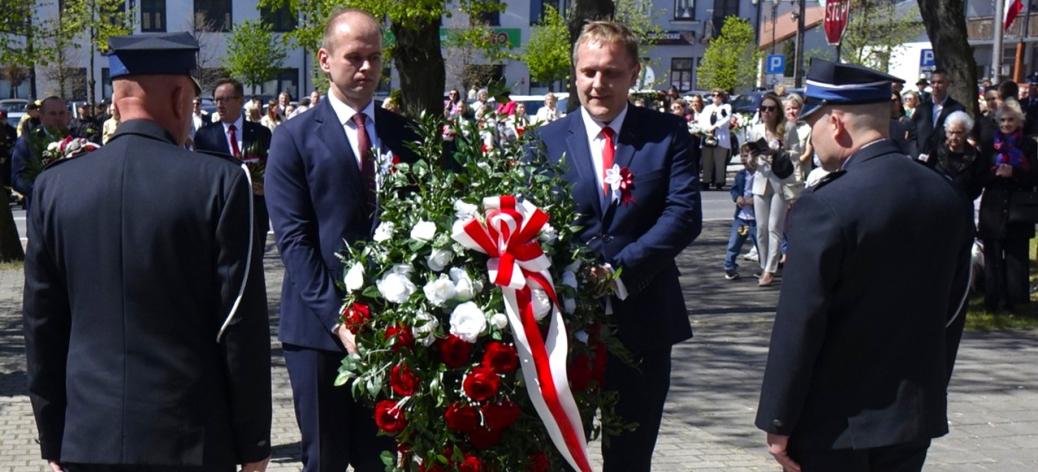 Dwóch urzędników w garniturach trzyma wieniec z biało-czerwonych kwiatów podczas uroczystości 3-majowych w parku na placu Wolności w Drzewicy. Obok stoją umundurowani strażacy, a w tle widoczny jest tłum uczestników oraz zabudowa miejska.