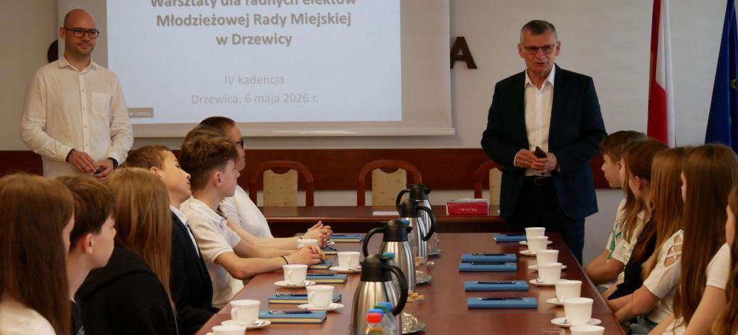 Na zdjęciu spotkanie warsztatowe: grupa młodzieży siedzi przy stole konferencyjnym, po prawej stronie stołu Burmistrza Drzewicy zabierający głos, po lewej Zastępca Burmistrza. Na ekranie wyświetlony jest slajd dotyczący warsztatów dla Młodzieżowej Rady Miejskiej w Drzewicy.