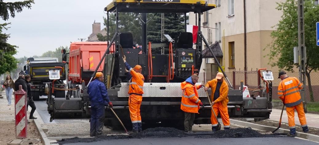 Prace drogowe na ulicy Braci Kobylańskich w terenie zabudowanym. Na środku duża maszyna do układania nawierzchni asfaltowej, za którą kilku pracowników w pomarańczowych strojach ochronnych rozprowadza świeżo ułożony asfalt. W tle samochody ciężarowe, budynki mieszkalne oraz drzewa. Pogoda pochmurna.