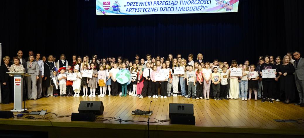  Na scenie sali widowiskowej RCK liczna grupa dzieci, młodzieży, opiekunów i samorządowców stoi w kilku rzędach przed ciemną kurtyną. Niektóre dzieci trzymają symboliczne czeki z nagrodami finansowymi oraz rekwizyty z wcześniejszych występów. Na górze duży biały transparent: „DRZEWICKI PRZEGLĄD TWÓRCZOŚCI ARTYSTYCZNEJ DZIECI I MŁODZIEŻY”. 