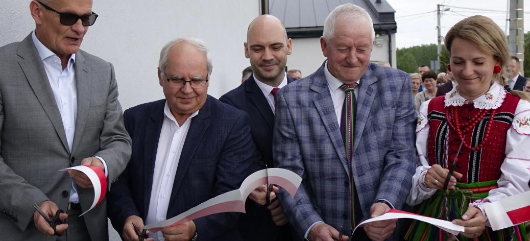  Zbliżenie na moment przecinania biało-czerwonej wstęgi na zewnątrz budynku. Wstęgę przecina pięć osób: Burmistrzowie Drzewicy, Prezes PKP Intercity Remtrak, Sołtys Idzikowic oraz Przewodnicząca KGW Idzikowice w tradycyjnym stroju ludowym.