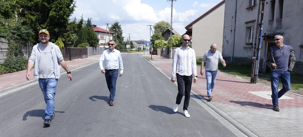 Pięciu mężczyzn idzie wzdłuż nowo wyremontowanej, asfaltowej ulicy w słoneczny dzień. Część idzie środkiem jezdni, pozostali po przylegającym do niej chodniku z kostki brukowej. W tle domy jednorodzinne, ogrodzenia oraz drzewa