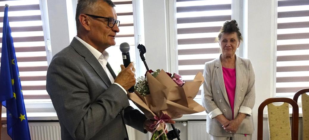 Burmistrz Drzewicy Janusz Reszelewski w szarym garniturze mówi do mikrofonu i przygotowuje się do wręczenia bukietu kwiatów stojącej naprzeciwko Skarbnik Gminy Irenie Jóźwickiej, ubranej w jasny garnitur i amarantową bluzkę.