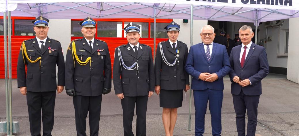 Grupa sześciu osób stojących przed budynkiem pod białym namiotem z napisem „Państwowa Straż Pożarna”. Cztery osoby ubrane są w galowe mundury strażackie, dwie pozostałe w garnitury. W tle znajdują się czerwone bramy garażowe jednostki straży pożarnej. Uczestnicy stoją w jednym rzędzie i pozują do wspólnego zdjęcia.