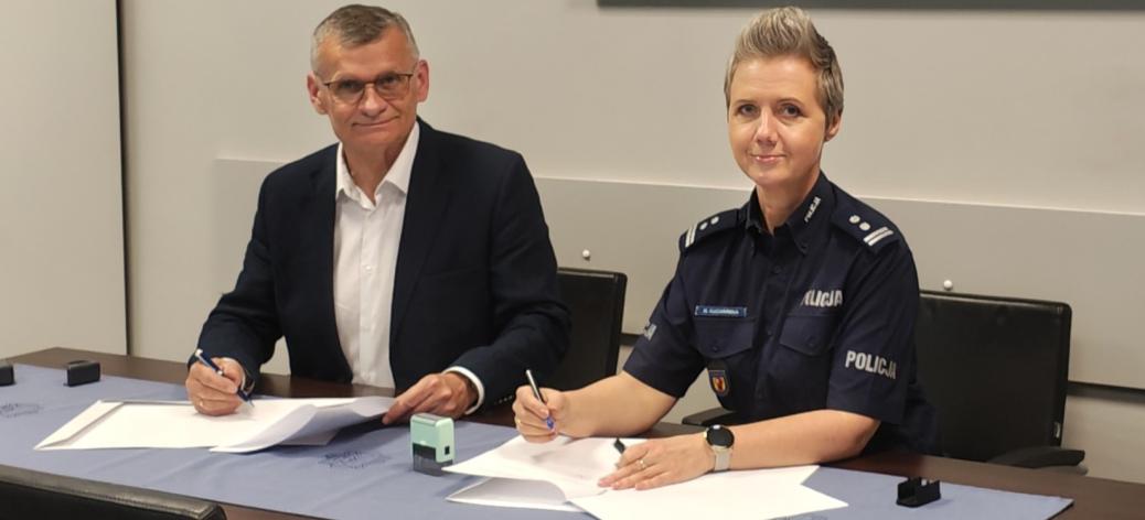  Podczas podpisywania porozumienia przy stole siedzą Burmistrz Drzewicy (w garniturze) i Pani Komendant Powiatowy Policji w Opocznie (w granatowym mundurze). Oboje trzymają długopisy i patrzą w stronę aparatu. Na stole znajdują się dokumenty oraz pieczątka. 