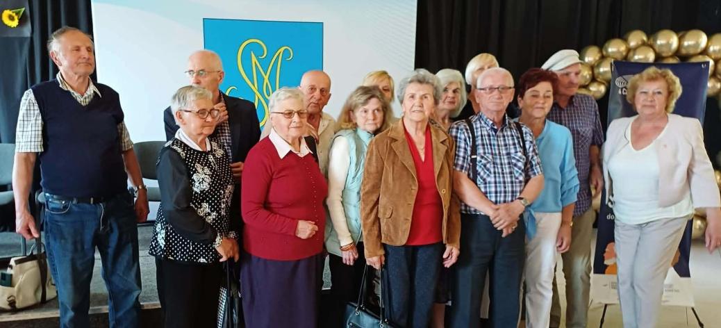 Grupowe zdjęcie kilkunastu seniorów, stojących blisko siebie w pomieszczeniu i pozujących do fotografii. Grupa składa się z kobiet i mężczyzn ubranych w stroje codzienne i wyjściowe. W tle czarna kotara, złote balony oraz jasna tablica z niebiesko-żółtym herbem przedstawiającym splecione litery. Po prawej stronie baner typu roll-up.