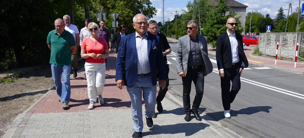 Oficjalna delegacja idzie w stronę obiektywu nowym chodnikiem z szarej kostki. W centrum I Wicewojewoda Łódzki w niebieskiej marynarce, otoczony przez innych uczestników spotkania.