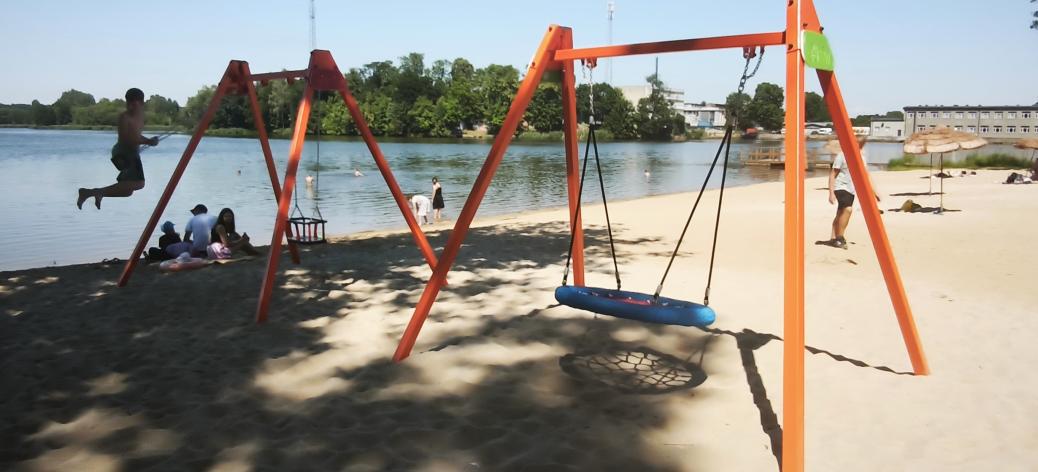 Nowoczesne huśtawki – tradycyjna oraz typu bocianie gniazdo – na pomarańczowych stelażach, ustawione na piaszczystej plaży nad jeziorem. W tle widoczni są kąpiący się i odpoczywający ludzie oraz zielony, przeciwległy brzeg.