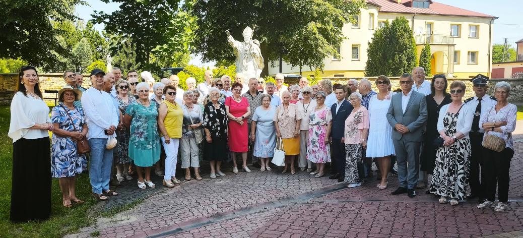 Liczna grupa uczestników spotkania pozuje do wspólnego zdjęcia na wybrukowanym placu kościelnym. W tle, za zebranymi ludźmi, widoczny jest biały pomnik przedstawiający papieża Jana Pawła II, otaczające go drzewa oraz żółty budynek mieszkalny.