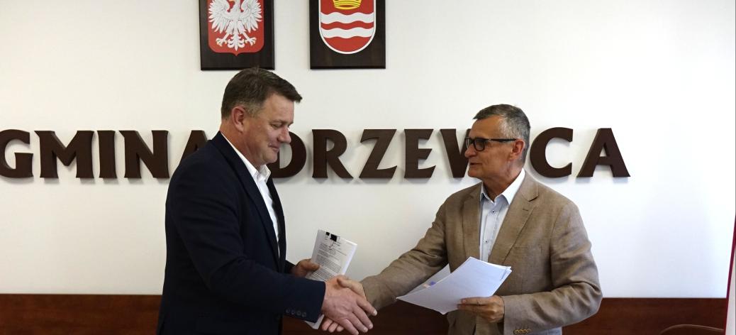 Członek Zarządu Woj. Łódzkiego Artur Bagieński i Burmistrz Drzewicy Janusz Reszelewski stoją w sali konferencyjnej i podają sobie dłonie w uścisku nad stołem, trzymając w rękach dokumenty. W tle biała ściana z dużym napisem „GMINA DRZEWICA”, nad którym wiszą godło Polski oraz herb Drzewicy.