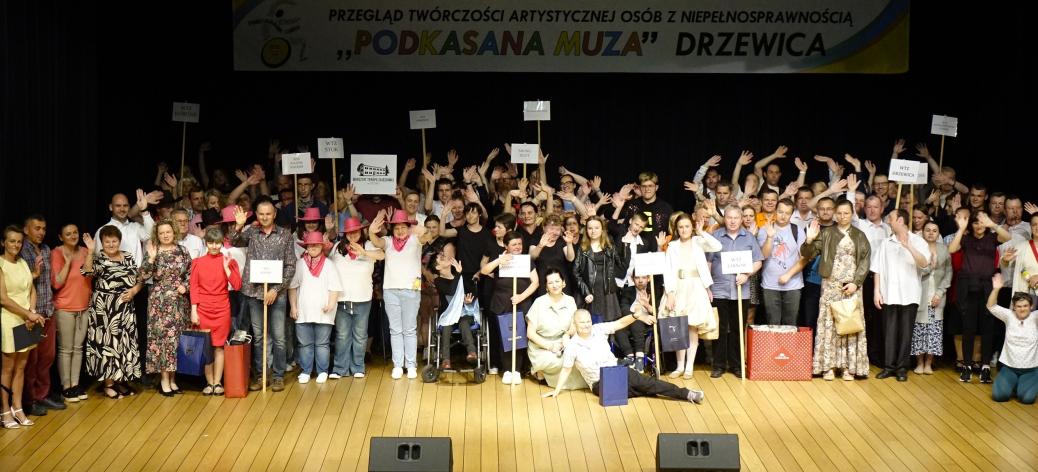 Na scenie w sali widowiskowej RCK kilkadziesiąt osób stoi obok siebie, uśmiechając się i machając rękami. W tłumie widoczne są osoby na wózkach inwalidzkich, osoby w różowych kapeluszach kowbojskich oraz trzymające tabliczki z nazwami różnych placówek (m.in. WTZ). Wysoko duży transparent festiwalu.