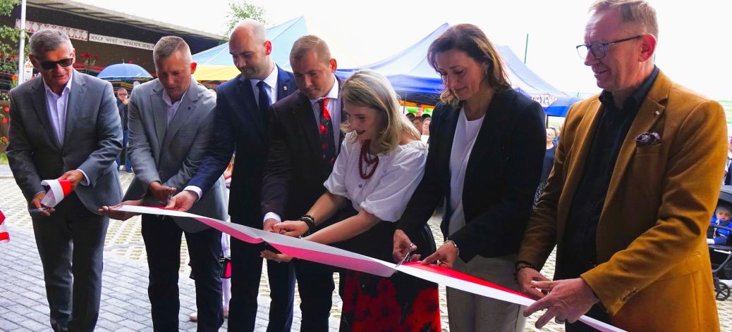 Oficjalny moment przecinania wstęgi. Grupa siedmiu osób – sześciu mężczyzn w garniturach lub marynarkach oraz dwie kobiety – stoi w rzędzie na zewnątrz budynku. Wspólnie trzymają i przecinają nożyczkami biało-czerwoną wstęgę. W tle widoczne są namioty festynowe oraz fragmenty otoczenia.