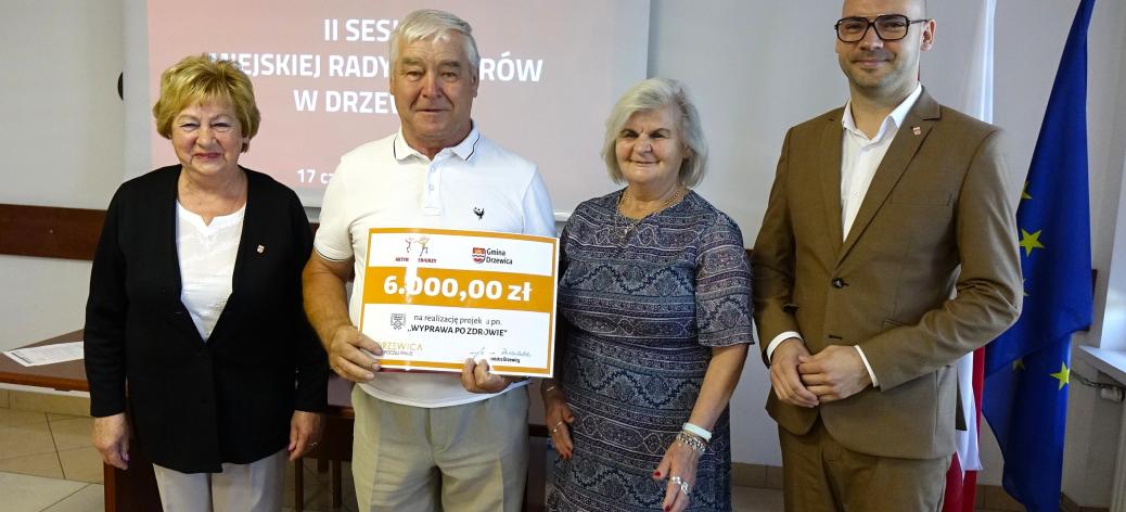 Pozowane zdjęcie czterech osób stojących obok siebie w sali konferencyjnej. Drugi od lewej mężczyzna trzyma duży, symboliczny czek na kwotę 6000,00 zł na realizację projektu „WYPRAWA PO ZDROWIE”. Po prawej stronie stoi mężczyzna w jasnobrązowym garniturze i okularach. W tle widoczny jest ekran projekcyjny z napisem „II SESJA MIEJSKIEJ RADY SENIORÓW W DRZEWICY” oraz flagi państwowe.