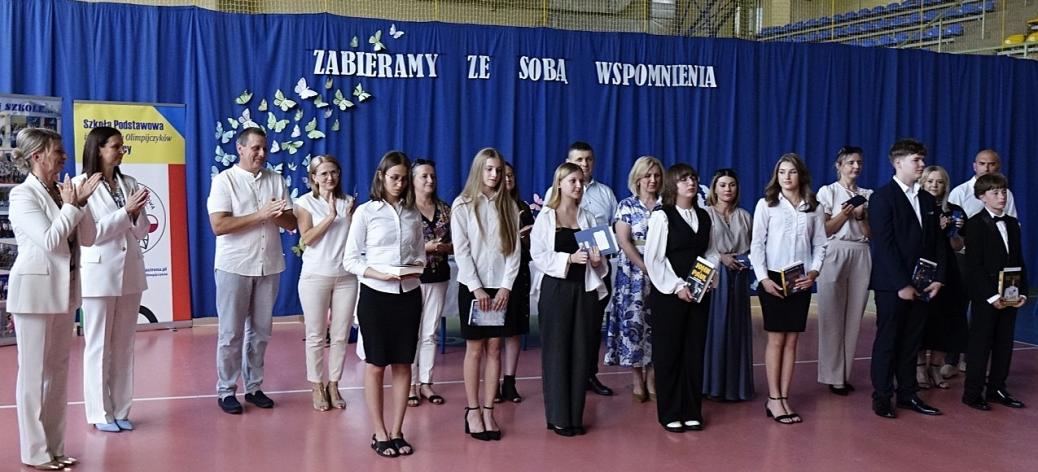 Uroczyste pożegnanie ósmoklasistów w Szkole Podstawowej im. Polskich Olimpijczyków w Drzewicy. Uczniowie w galowych strojach odbierają nagrody książkowe na tle niebieskiej kurtyny z napisem "ZABIERAMY ZE SOBĄ WSPOMNIENIA"