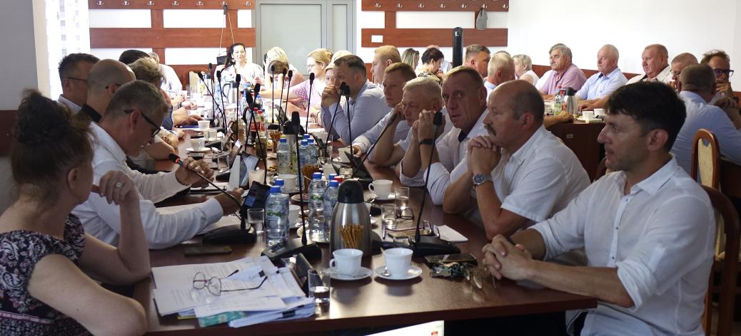 Szeroka panorama sali konferencyjnej Urzędu Miejskiego w Drzewicy. Przy długim stole siedzi kilkunastu mężczyzn i kobiet ubranych oficjalnie. Uczestnicy sesji, w tym Burmistrz Janusz Reszelewski, skupieni są na dokumentach i ekranie, na którym widoczny jest porządek obrad XXV sesji Rady Miejskiej. Na stole znajdują się mikrofony, butelki z wodą i filiżanki.