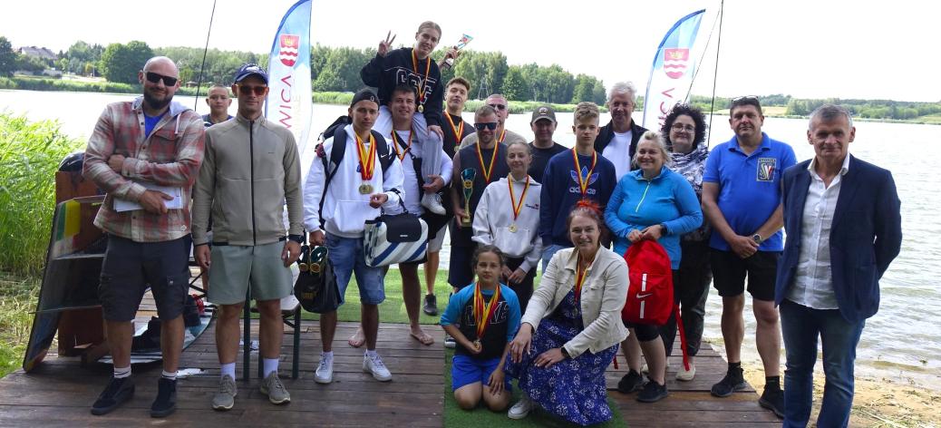 Grupa kilkunastu osób w różnym wieku (młodzież i dorośli) pozuje do wspólnego zdjęcia na drewnianym pomoście nad wodą. Część osób ma na szyi medale, a w tle widać flagi reklamowe z herbem Drzewicy. Wszyscy są w letnich, sportowych strojach. Na pierwszym planie po prawej stronie stoi Burmistrz Drzewicy w ciemnej marynarce i jasnej koszuli, a obok niego, na środku pomostu, klęczy kobieta w niebieskiej spódnicy w kwiaty oraz młoda dziewczynka w sportowym stroju.