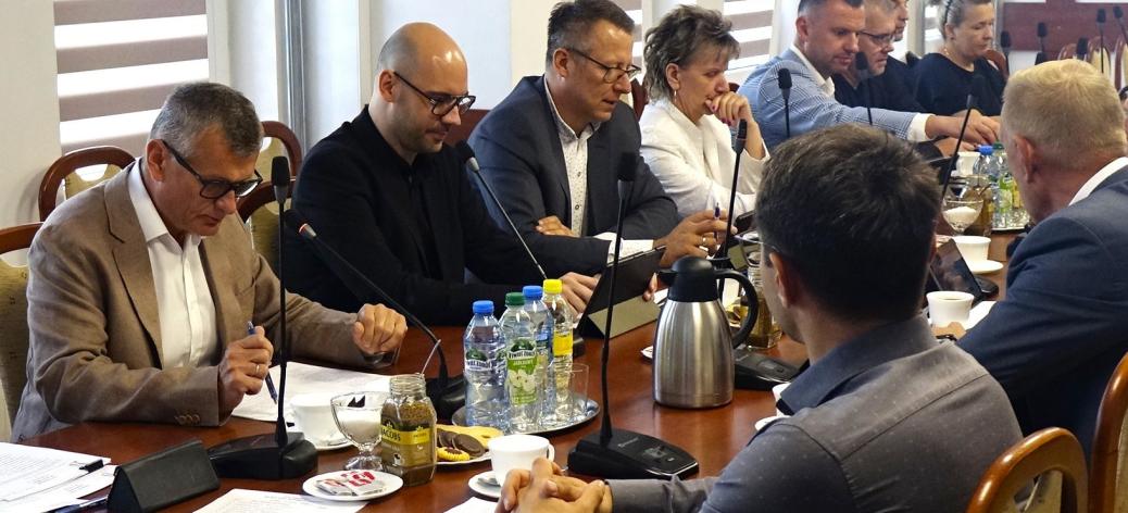 Uczestnicy sesji Rady Miejskiej w Drzewicy siedzą po obu stronach stołu konferencyjnego i biorą udział w obradach. Pierwszy z lewej Burmistrz Drzewicy odczytuje dokument. Na stołach znajdują się dokumenty, mikrofony oraz napoje.
