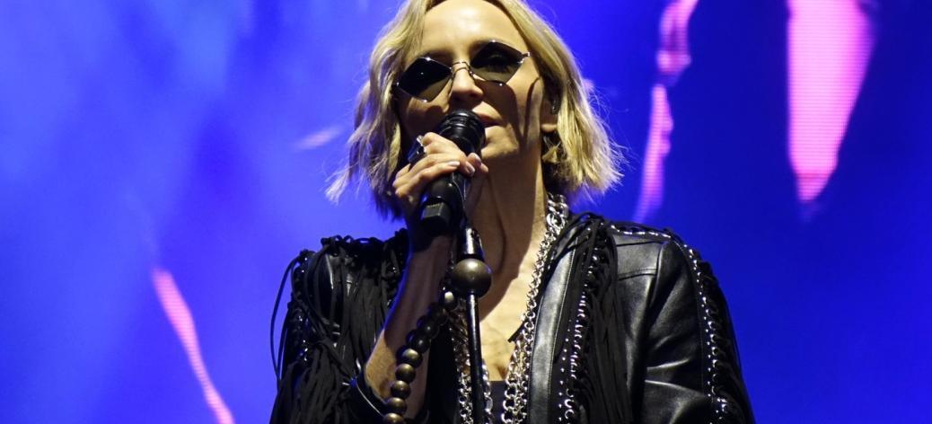 Anna Wyszkoni podczas koncertu śpiewa do mikrofonu na oświetlonej niebiesko-fioletowym światłem scenie. Artystka ma krótkie blond włosy, ciemne okulary i czarną skórzaną kurtkę z frędzlami. W tle widoczny jest ekran LED