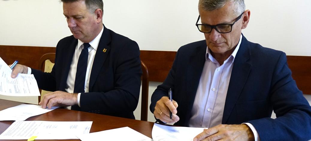 Członek Zarządu Woj. Łódzkiego Artur Bagieński i Burmistrz Drzewicy Janusz Reszelewski w garniturach siedzą obok siebie przy drewnianym konferencyjnym stole. Obaj podpisują leżące przed nimi dokumenty. Na ścianie w tle widoczny duży, brązowy, wypukły napis „GMINA DRZEWICA”.