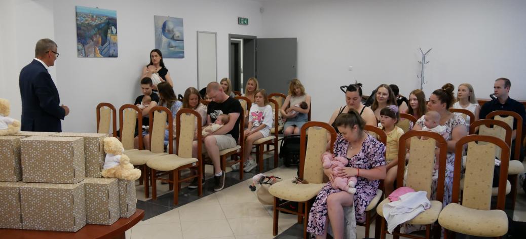 Burmistrz Drzewicy przemawia do rodzin zgromadzonych w jasnej sali urzędowej. Po lewej stronie, na stole, ułożone są przygotowane upominki w ozdobnych pudełkach z pluszowymi misiami. W rzędach krzeseł siedzą rodzice trzymający na rękach noworodki i małe dzieci.