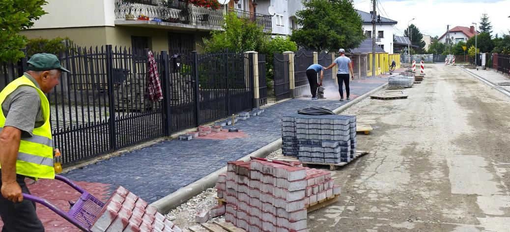 a osiedlowej ulicy trwają prace związane z przebudową drogi. Widoczne są nowo ułożone chodniki z kostki brukowej, krawężniki, materiały budowlane oraz pracownicy wykonujący roboty. W tle znajdują się domy jednorodzinne.