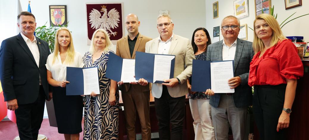 Grupa ośmiu osób w gabinecie pozuje do wspólnego zdjęcia. Cztery osoby prezentują otwarte niebieskie teczki z podpisanymi dokumentami. W środku Zastępca Burmistrza Drzewicy Dominik Niemirski.