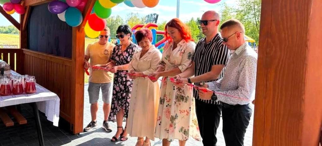 roczyste przecięcie czerwonej wstęgi pod drewnianą wiatą w Domasznie, przyozdobioną kolorowymi balonami podczas Pikniku Rodzinnego.