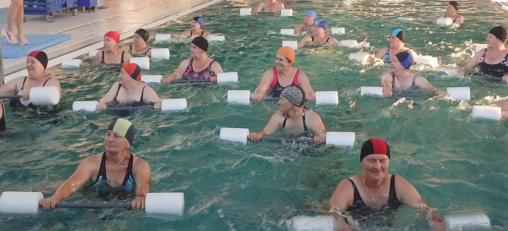 Liczna grupa seniorek wykonująca ćwiczenia w basenie z użyciem piankowego sprzętu do aqua aerobiku.
