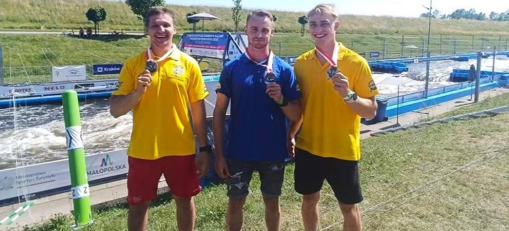 Trzej uśmiechnięci kajakarze górscy LKK Drzewica - od lewej: Szymon Zawadzki, Michał Franczak, Maciej Bartos - stoją na trawie przy torze kajakowym i prezentują zdobyte medale. Dwaj z nich mają na sobie żółte koszulki polo, a środkowy – niebieską koszulkę polo.