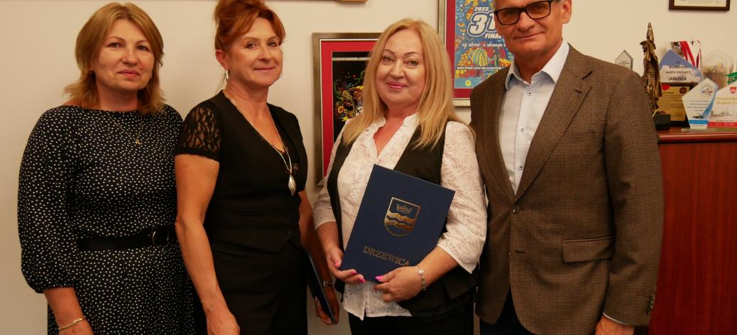 Pozowane zdjęcie w gabinecie Burmistrza Drzewicy. Od lewej trzy panie -  Dyrektor ZEAS, Dyrektor SP w Domasznie, Dyrektor SP w Domasznie – oraz Burmistrz Drzewicy. Na ścianie za nimi wiszą oprawione plakaty 31. i 32. Finału WOŚP oraz rzeźbiona drewniana tablica.
