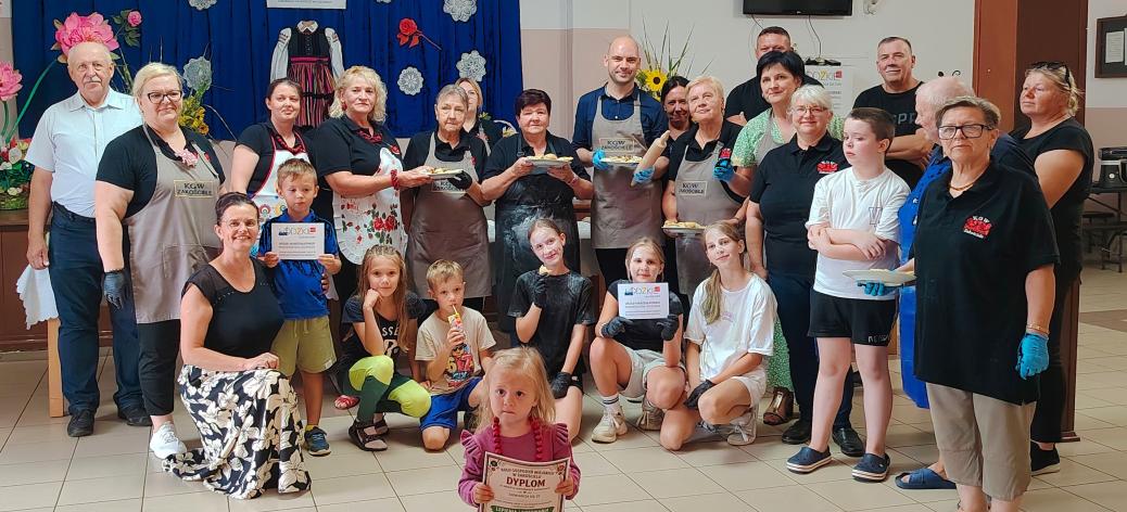  Uczestnicy i organizatorzy warsztatów lepienia pierogów pozują do pamiątkowego zdjęcia na świetlicy wiejskiej w Zakościelu. Część osób trzyma dyplomy i talerze z gotowymi pierogami, a na pierwszym planie mała dziewczynka dumna prezentuje oprawiony dyplom za udział w warsztatach.