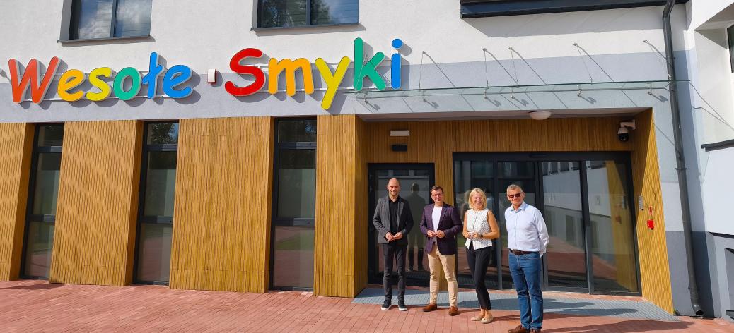 Cztery osoby stoją przed nowoczesnym budynkiem z kolorowym napisem „Wesołe Smyki” nad wejściem.