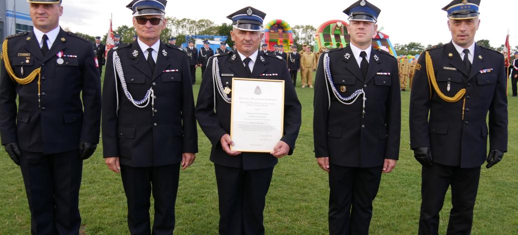Pięciu strażaków - Komendant Powiatowy PSP w Opocznie, Prezes Oddziału M-G ZOSP w Drzewicy (Burmistrz), Prezes OSP Radzice, Naczelnik OSP Radzice i Zastępca Komendanta Wojewódzkiego PSP w Łodzi - w galowych mundurach pozuje na boisku. W środku Prezes OSP Radzice trzyma oprawiony akt włączenia jednostki do KSRG. 