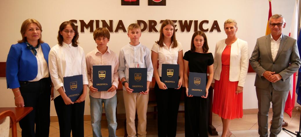 Pozowane zdjęcie grupowe w sali konferencyjnej Urzędu Miejskiego w Drzewicy. Pięcioro uczniów trzymających niebieskie pamiątkowe teczki stoi w centrum. Po bokach Pani Dyrektor ZEAS i Burmistrz Drzewicy. W tle widoczny napis „GMINA DRZEWICA” oraz godło i herb.