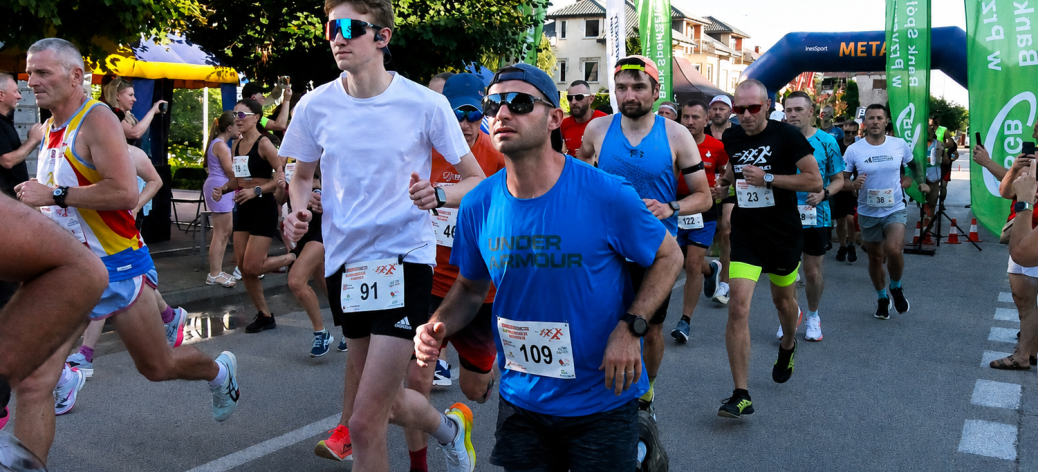 Grupa biegaczy w trakcie wyścigu ulicznego Drzewica Summer Run. Na pierwszym planie mężczyzna w niebieskiej koszulce z numerem 109 i okularach przeciwsłonecznych, obok biegacz w białej koszulce.