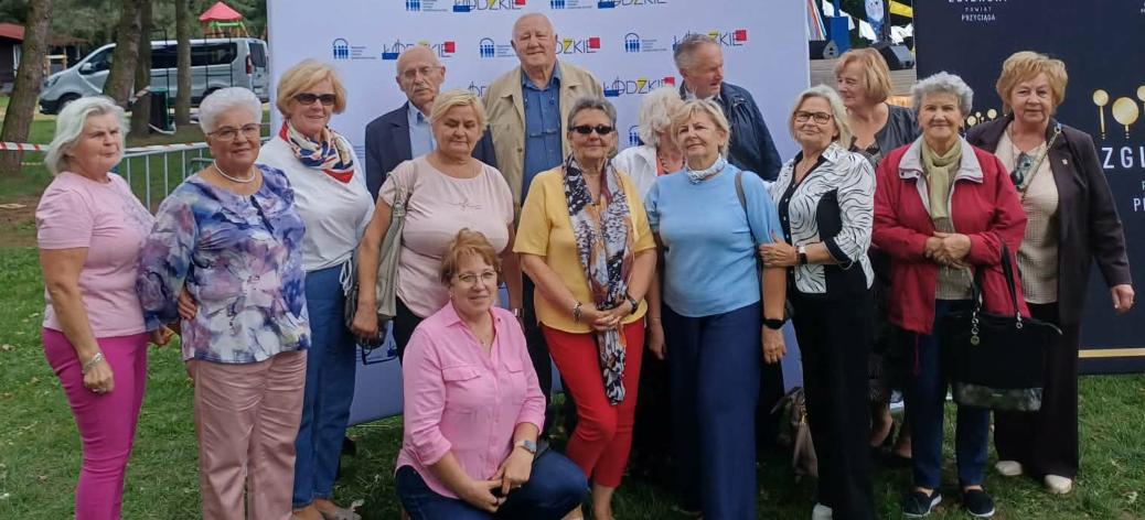 Grupa kilkunastu seniorów reprezentujących Gminę Drzewica pozuje do pamiątkowej fotografii w plenerze na tle promocyjnej ścianki Województwa Łódzkiego podczas Wojewódzkich Obchodów Dnia Seniora