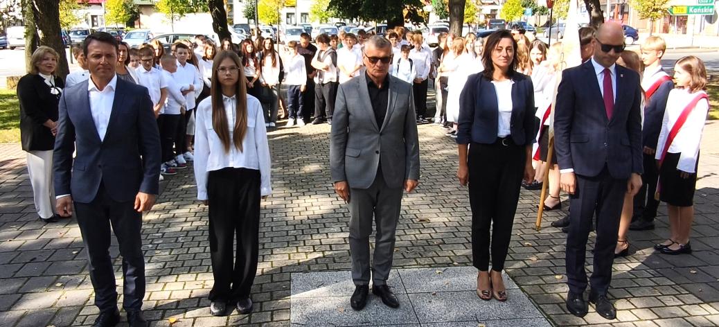  Pięć osób w strojach galowych – samorządowcy i uczennica SP w Drzewicy - stoi w szeregu w parku wyłożonym kostką przed Pomnikiem Pamięci w Drzewicy. W tle widać zgromadzoną młodzież szkolną, poczet sztandarowy i drzewa.