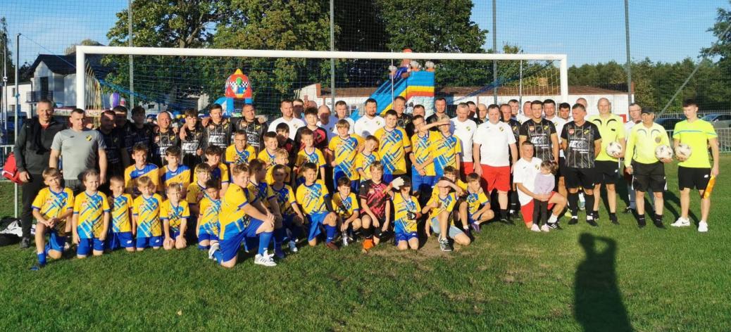 Grupowe zdjęcie zawodników, młodych piłkarzy oraz sędziów na boisku przed bramką.