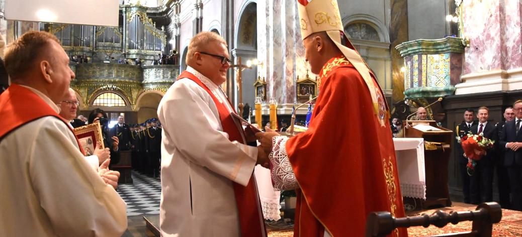 Uroczystość kościelna we wnętrzu świątyni w Studziannie. Biskup Marek Solarczyk w czerwonej szacie liturgicznej i mitrze wręcza akt nominacji ks. Grzegorzowi Włodarczykowi w białej albie i czerwonej stule. Z lewej strony przygląda się inny duchowny. W tle zabytkowe organy, nawy kościoła oraz zgromadzeni wierni i strażacy. 