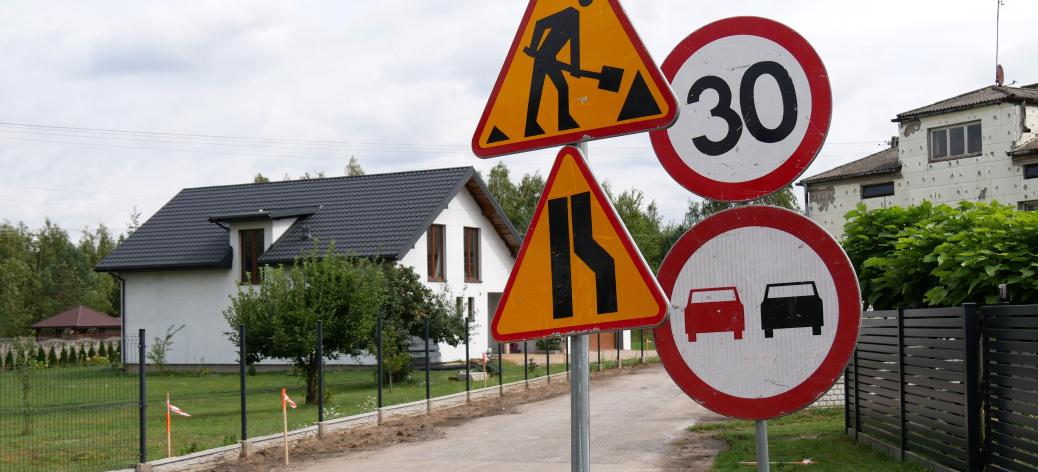 Znaki drogowe ostrzegające o robotach drogowych, zwężeniu jezdni, ograniczeniu prędkości do 30 km/h oraz zakazie wyprzedzania, ustawione przy remontowanej drodze w sąsiedztwie domów jednorodzinnych.
