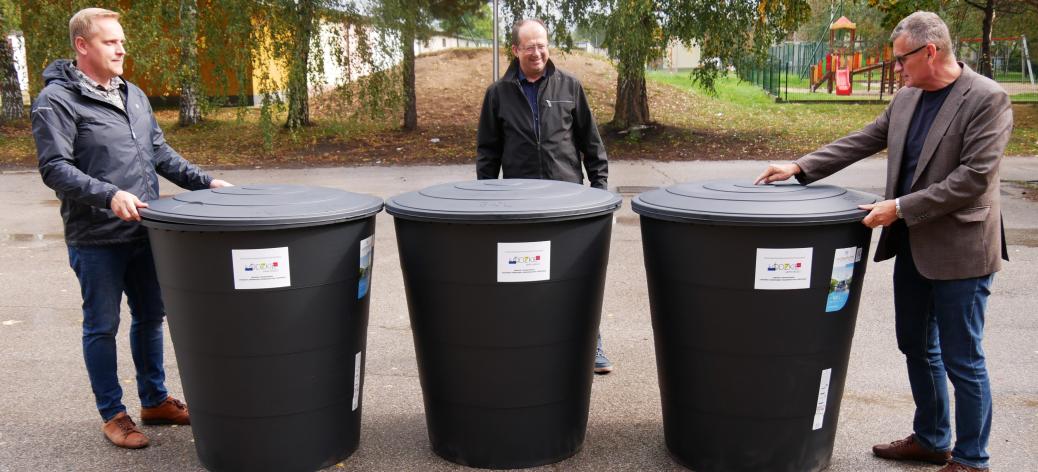  Burmistrz Drzewicy i pracownicy UM w Drzewicy - stoi na placu obok trzech dużych, czarnych pojemników na deszczówkę oklejonych informacyjnymi naklejkami.