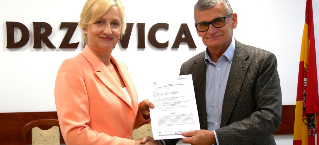 Pani Dyrektor SP w Radzicach w łososiowym garniturze oraz Burmistrz Drzewicy w okularach i szarej marynarce uściskają sobie dłonie, wspólnie trzymając podpisane umowy. W tle ściana z napisem „Drzewica”, godło i herb oraz fragment flagi.