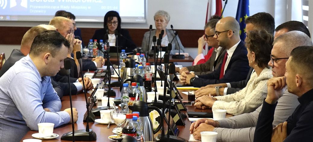 Sala konferencyjna UM w Drzewicy, w której odbywa się posiedzenie. Przy długim stole, na którym ustawiono mikrofony, filiżanki, butelki z napojami oraz dzbanki, siedzą po obu stronach radni i uczestnicy obrad. Na dalszym planie, na przedzie sali, zasiadają członkowie Prezydium Rady Miejskiej. Za nimi ekran projekcyjny oraz flagi.   