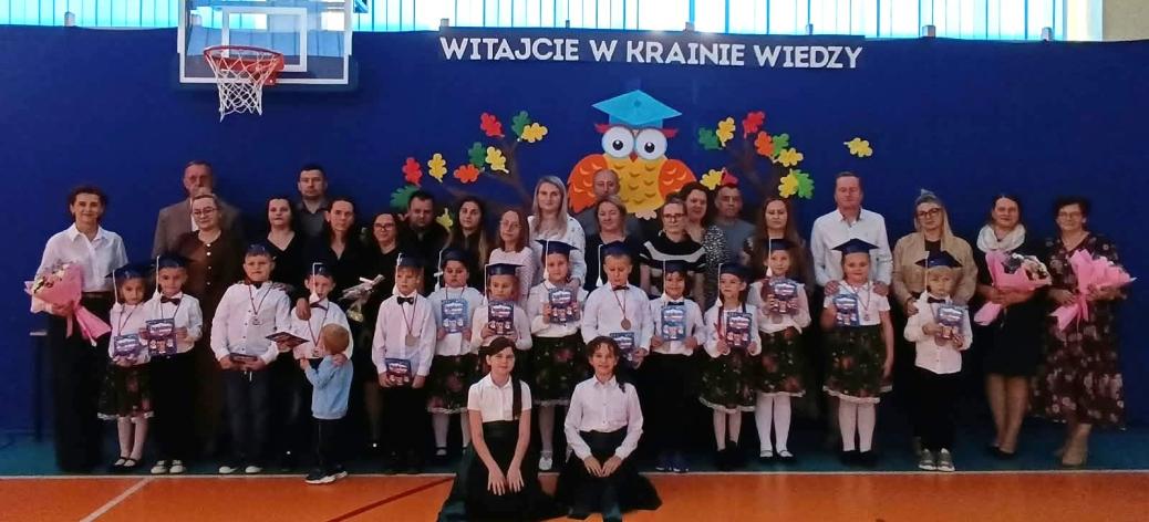 Grupowo pozujące dzieci w galowych strojach, nauczyciele i rodzice podczas uroczystości szkolnej w sali gimnastycznej SP w Brzustowcu. W tle niebieska ściana z napisem „WITAJCIE W KRAINIE WIEDZY” oraz dekoracją z sową i jesiennymi liśćmi.