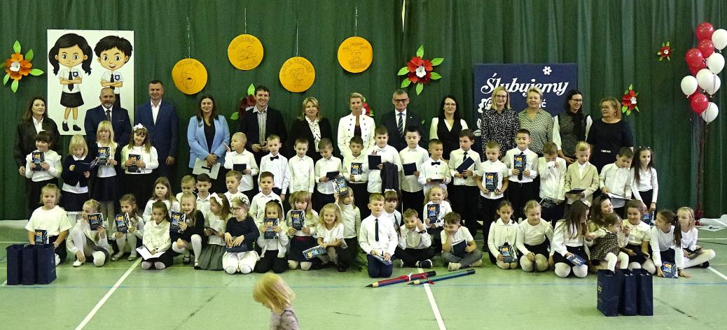 Grupowa fotografia uczestników uroczystości na tle zielonej kotary udekorowanej papierowymi kwiatami, godłem, dekoracyjnymi kołami oraz napisem „Ślubujemy”. W centrum duża grupa dzieci w galowych strojach stojących oraz siedzących na podłodze, z których część trzyma w rękach pamiątkowe dyplomy lub książki. Za dziećmi stoją nauczyciele i zaproszeni goście. 